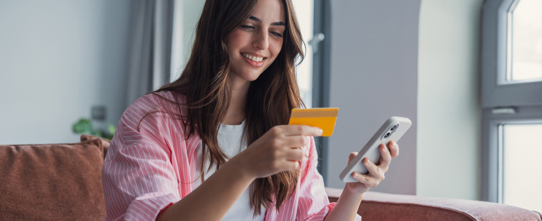 : Mujer sonriente sentada sosteniendo una tarjeta de crédito dorada y mirando su teléfono móvil de forma alegre.